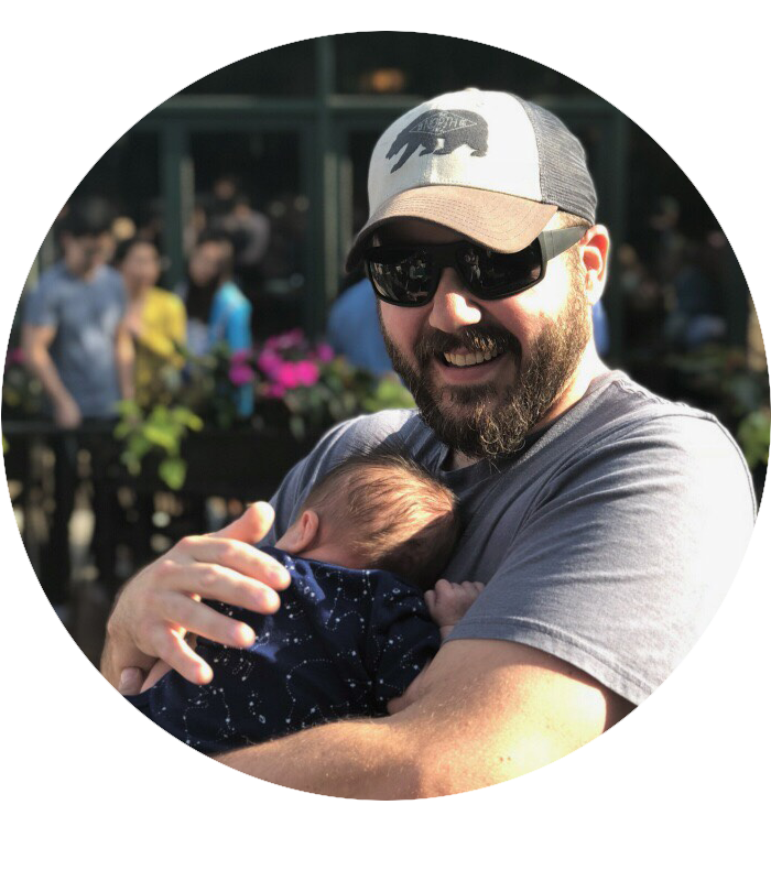 A picture of Justin. He has a beard, is wearing a baseball cap and sunglasses and is cradling his infant son.